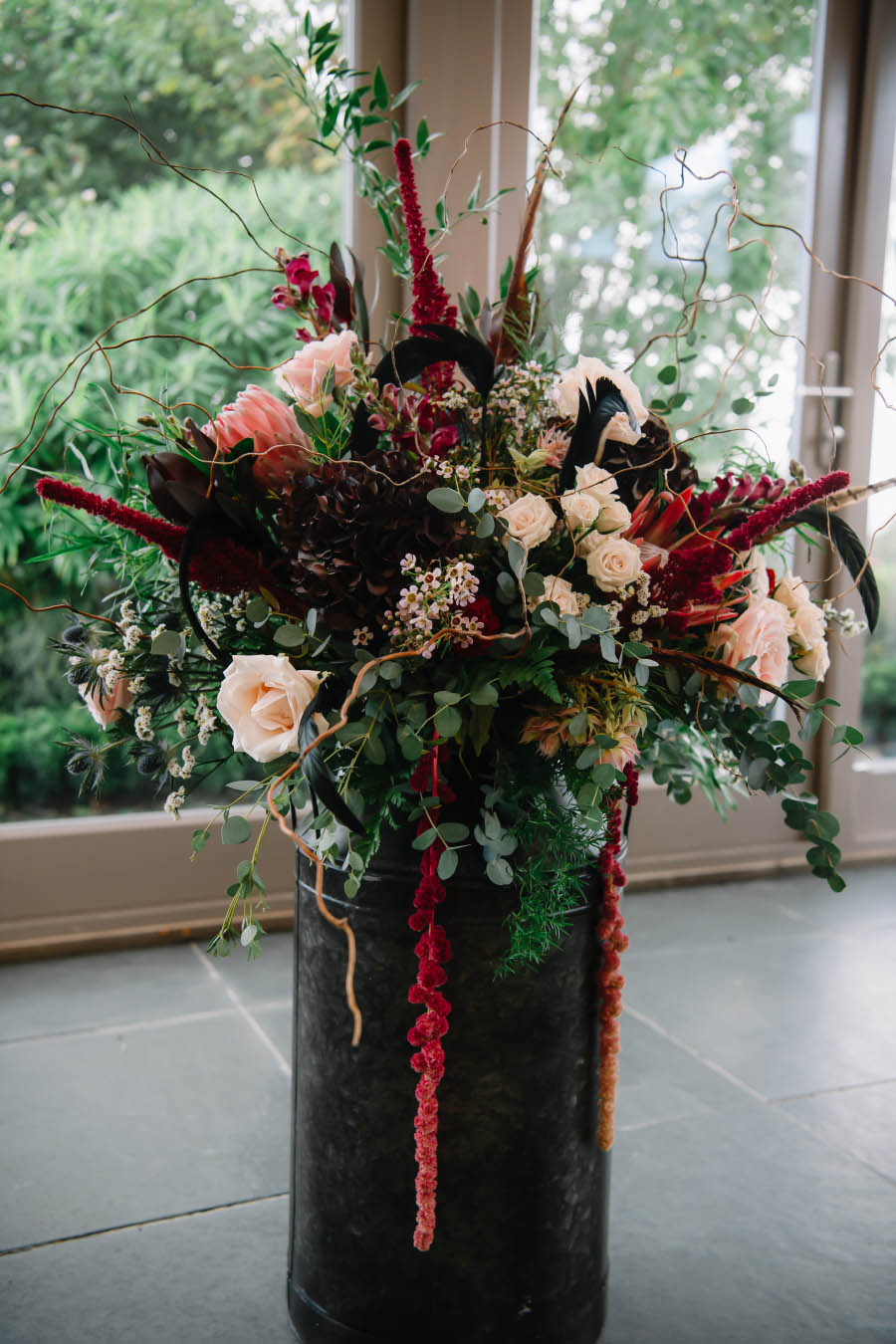 A black milk churn bears a rich bouquet of wedding flowers featuring roses and protea at a Gothic-inspired wedding at Trevenna Barns Cornwall, with wedding flowers by Artem Florum.