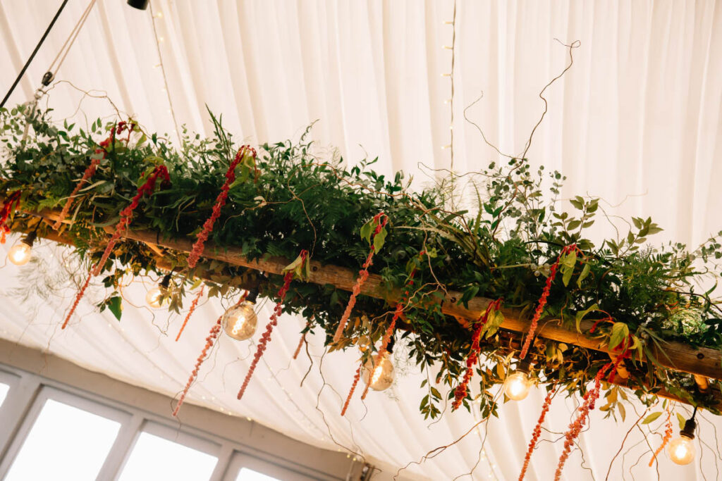 A suspended hanging ladder, artistically adorned with twisted hazel and rich red amaranths oversee the wedding banquet at a Gothic-inspired wedding at Trevenna Barns Cornwall. Wedding flowers by Artem Florum.